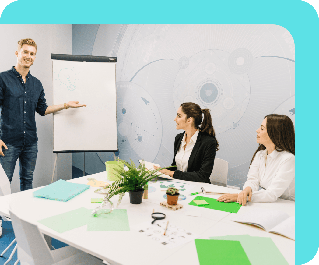 A business meeting in a bright office with a man explaining concepts to two attentive women at a white table, surrounded by documents and office supplies.